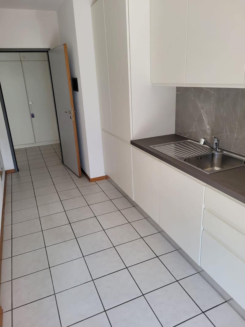 Compact kitchen with white cabinets, gray countertop, tiled floor, and a stainless steel sink with a dishwasher visible.