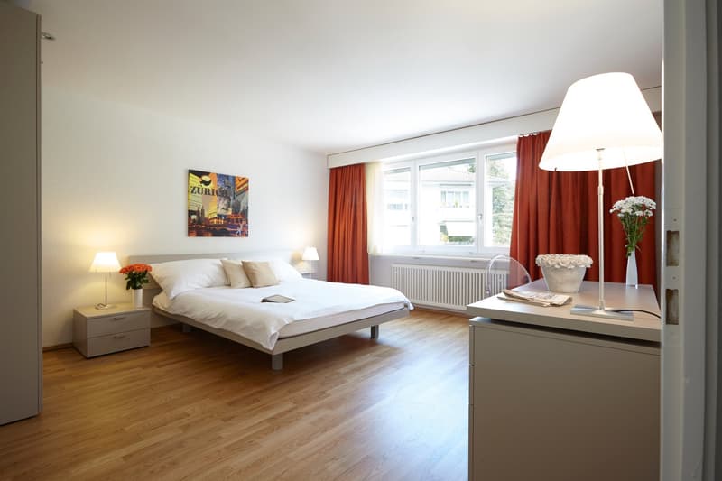 Spacious bedroom with hardwood flooring, large windows, white walls, a double bed, bedside tables, a dresser, and a floor lamp. The room also features red curtains and a framed artwork on the wall.
