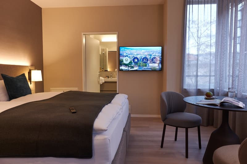 a bedroom with a bed, a remote control, a nightstand, a wall-mounted TV, a chair, a round table, a curtain, a pillow, a bathroom with a sink, and a lamp
