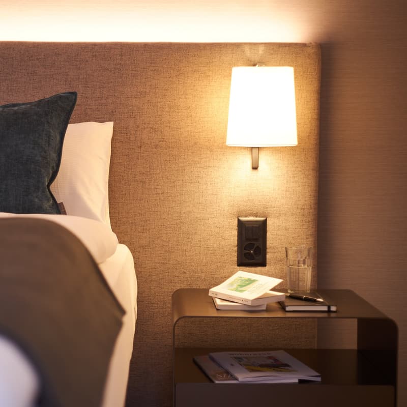 a bed with pillows, nightstand with books and pen, wall-mounted lamp