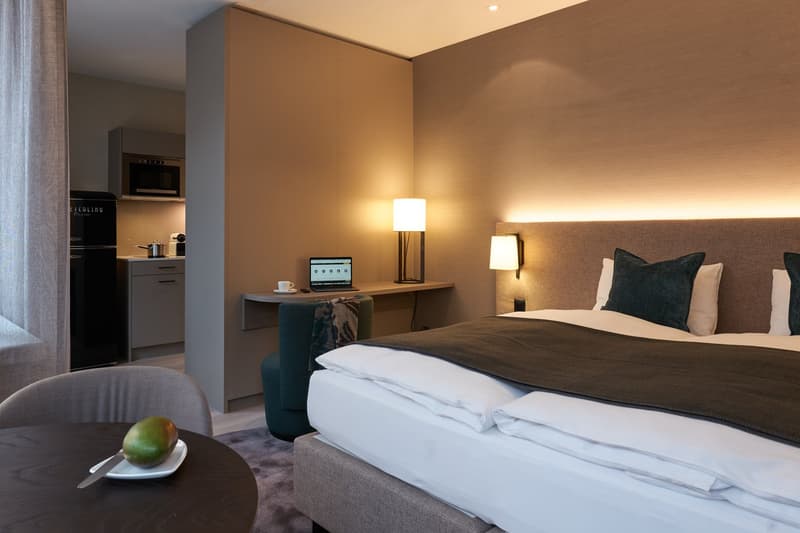 Double bed, bedspread, pillows, lamps, desk, laptop, chair, table, refrigerator, microwave, kettle, kitchen cabinet, grey walls