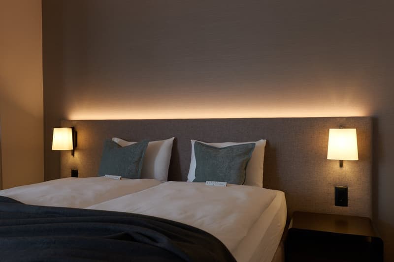 Double bed with white bedding and blue pillows, bedside lamps on both sides, gray headboard, small nightstand