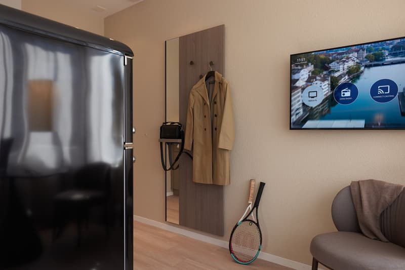 A room with a black refrigerator, coat rack with a beige trench coat, black handbag, a mounted television displaying a scenery, a tennis racket, and a grey armchair with a brown towel