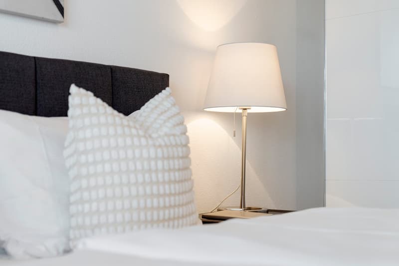 A modern, minimalistic bedroom with white walls and tile. A white bed and two white pillows are visible, along with a lamp, table, and nightstand.