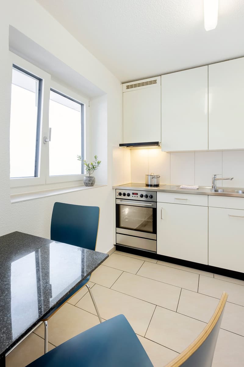 Open kitchen with integrated stove, oven, and sink, white tiles and cabinets, modern design, blue chairs, granite countertop, open window
