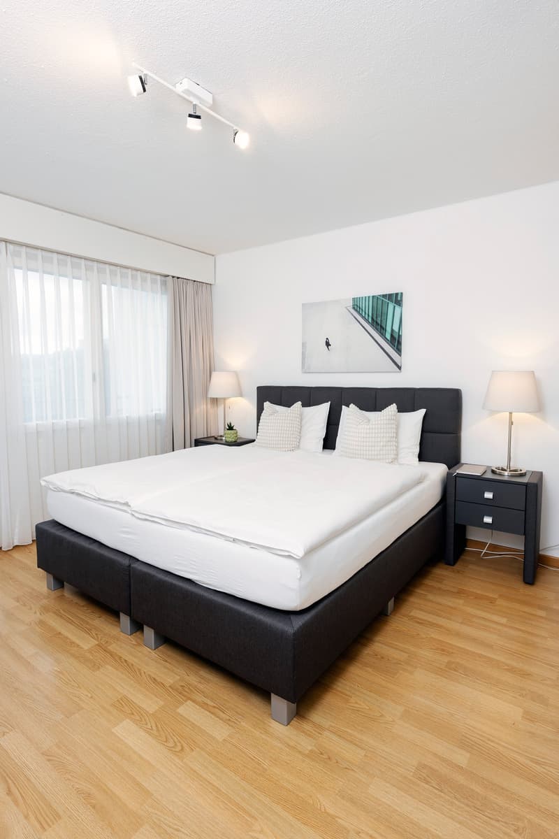 spacious bedroom with large bed, lamps, side tables, nightstands, white linens, framed photo, wooden floor, large window with white curtains
