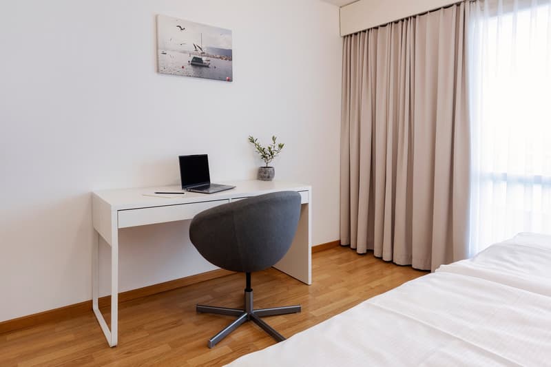 A white desk with a laptop, a plant, and a chair. An empty bed is present on the right side.