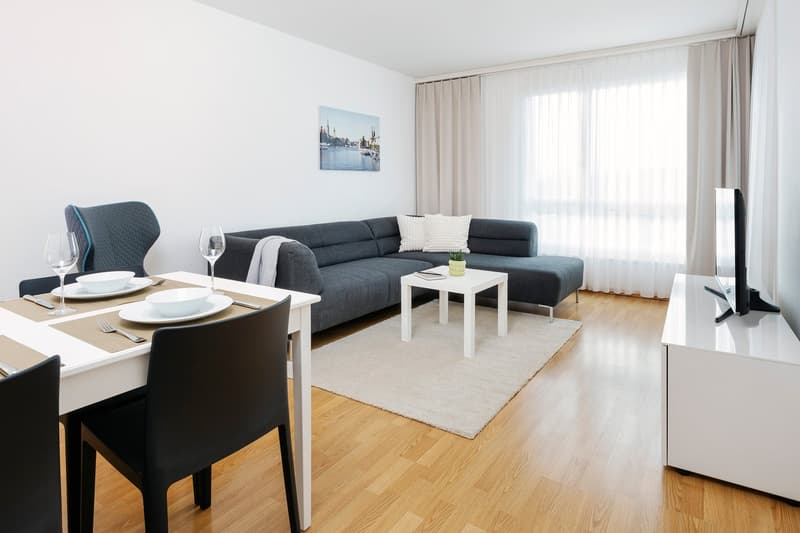 Living area with a sectional sofa, dining table, white wooden floor, large windows with curtains, wall art, small table with plant, flat screen television