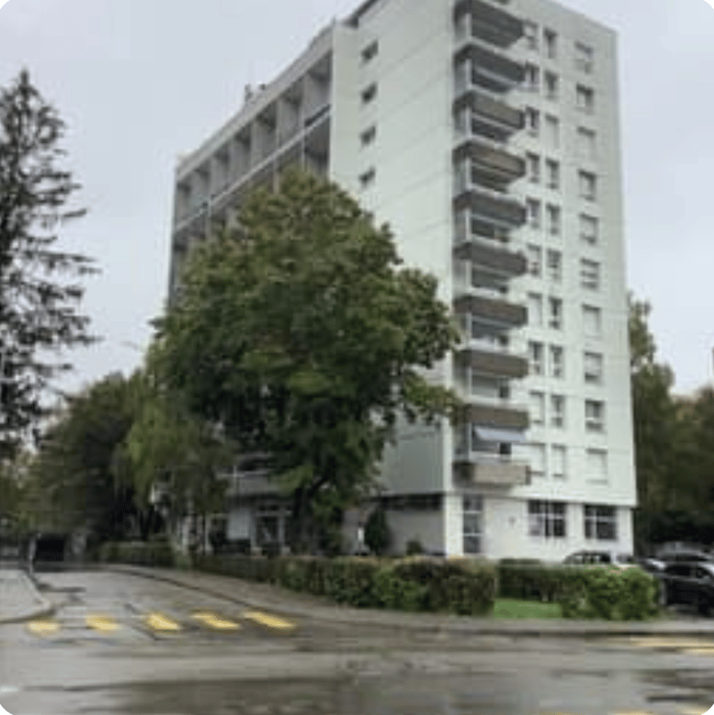 High-rise building, white exterior, multiple balconies, trees, shrubs, street