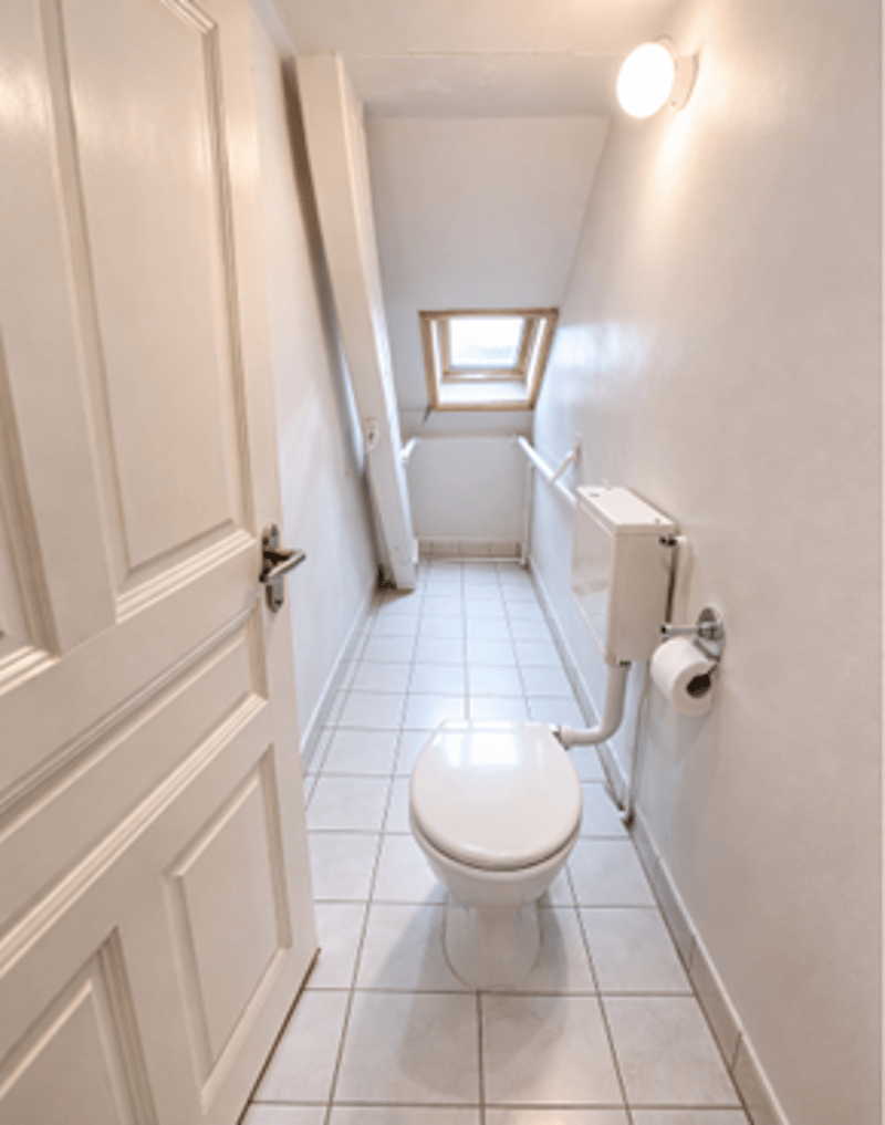 A small bathroom with white tiles, a toilet, a roll of toilet paper, a small window, and a light on the ceiling.