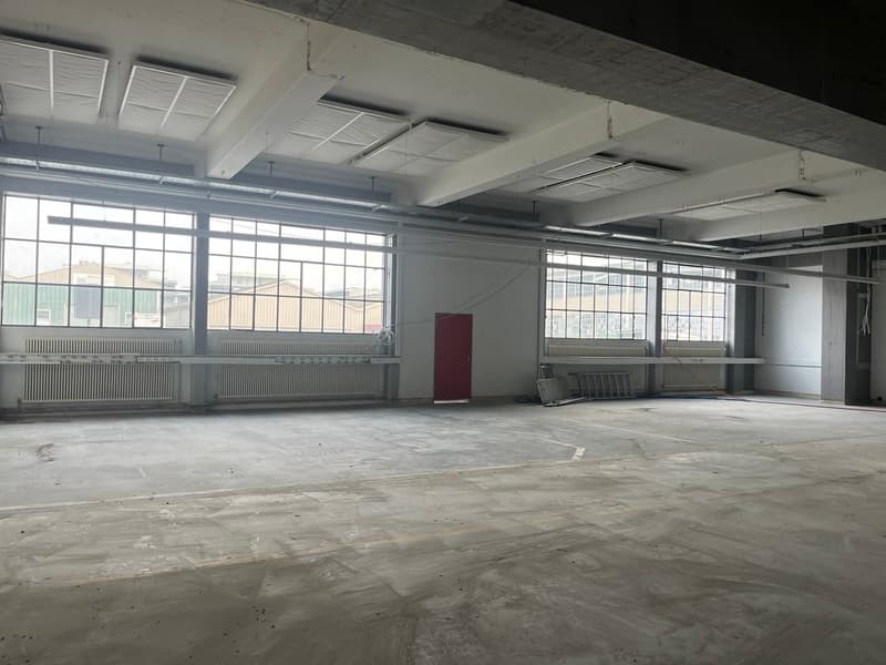 Large open industrial space with concrete floors, exposed ceiling beams, and large windows providing natural light. The space appears to be in a state of renovation or construction, with some equipment and materials visible.