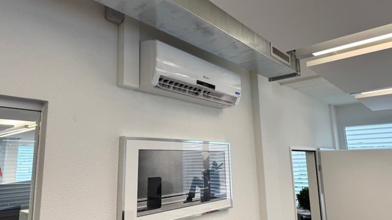 The image shows an office space with a white wall, a mounted flat-screen TV, and an air conditioning unit installed on the wall.