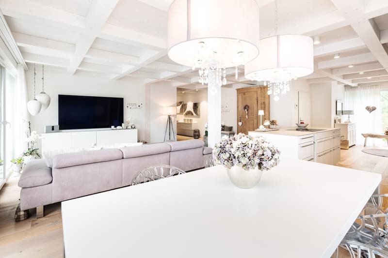 modern living room with white walls, wooden floor, large windows, white dining table, gray sofa, chandeliers, TV, kitchen with white cabinets, island with sink, flowers in vase