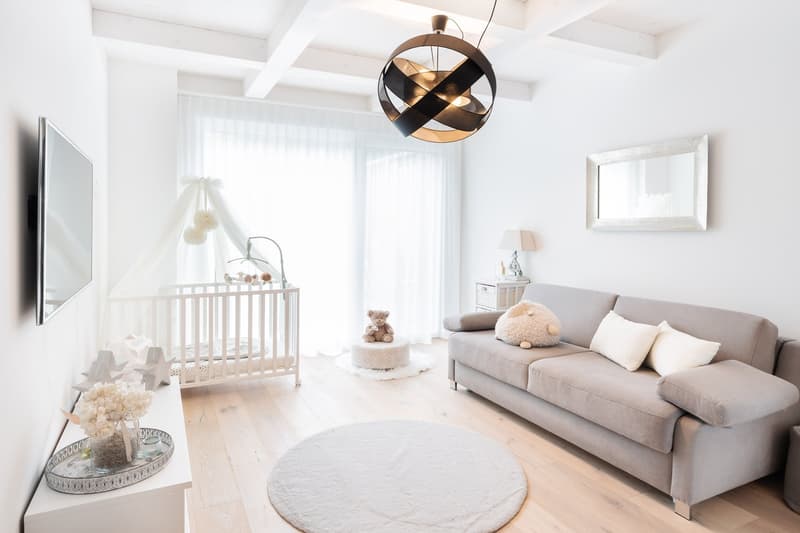 living room with white walls, wooden floor, gray couch, round rug, crib, TV, mirror, lamp, decorative items