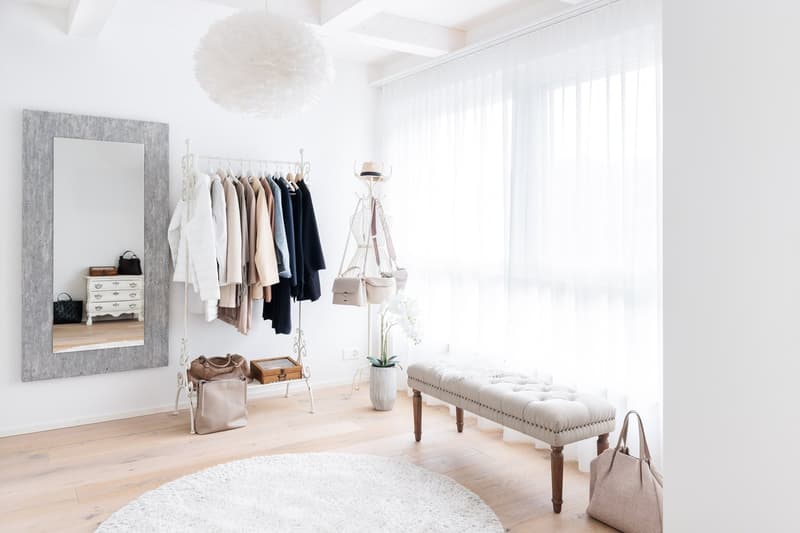 white walls, wooden floor, large mirror, clothes rack, bench, round rug, white curtains