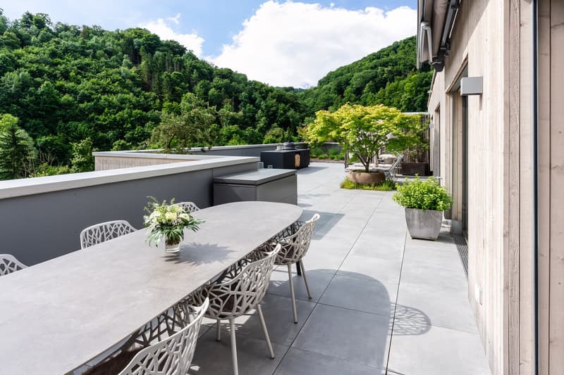 Outdoor dining area with a long table, chairs, and a flower vase. Potted plants and a grill are also present. The area has a view of the surrounding trees and mountains.