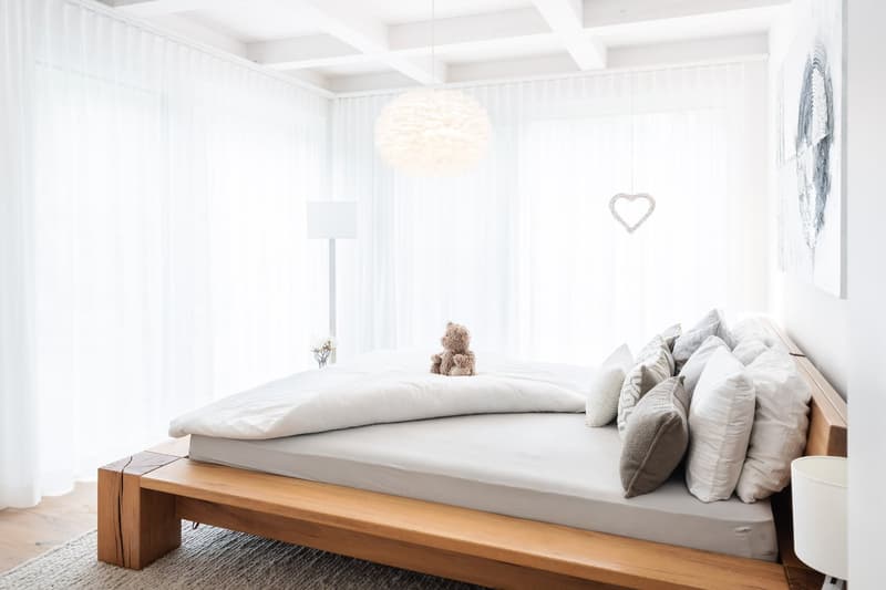 A bedroom with a wooden bed, white bedding, pillows, a teddy bear, white curtains, a white lamp, and a white heart decoration.