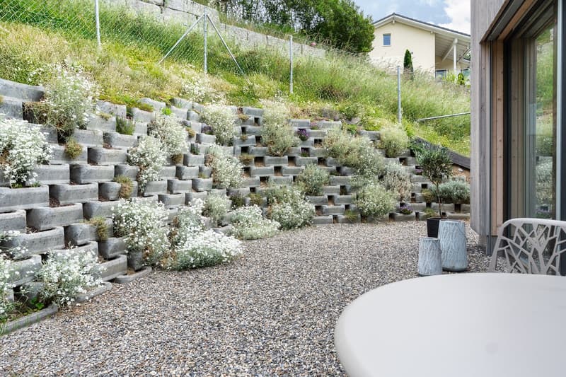 gravel pathway, concrete retaining wall with plants, white table and chair, potted plants