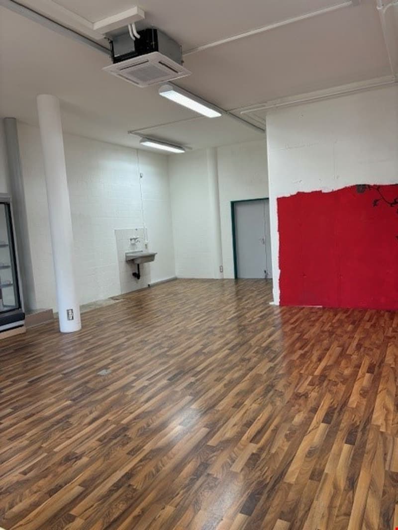 The image shows a large open office space with white walls, a hardwood floor, and a red accent wall. The room is equipped with overhead lighting and an air conditioning unit. The space appears to be suitable for use as an office or studio.