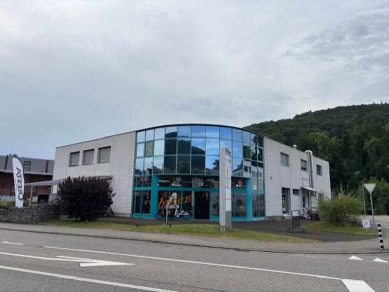 A modern, multi-story commercial building with a glass facade and a parking area in front of it. The building appears to be located near a road or highway.