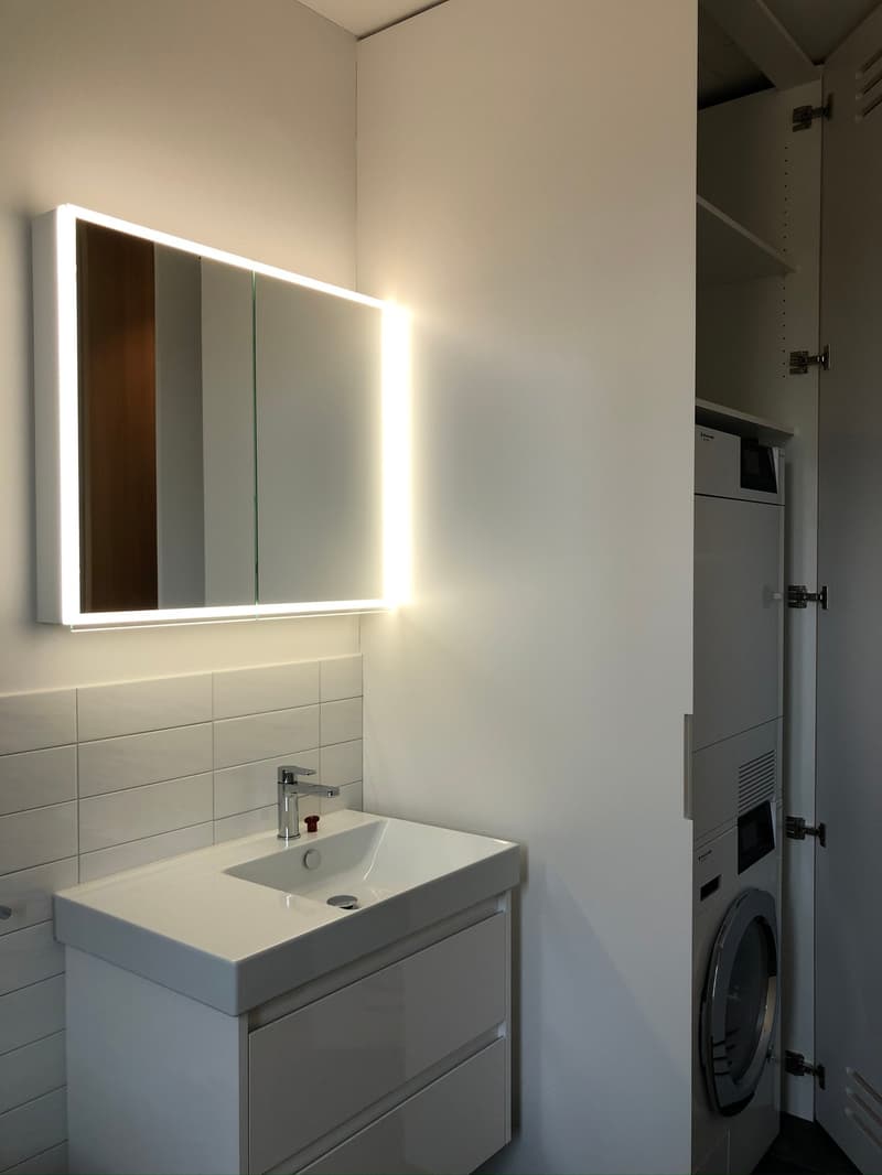 Bathroom with white sink, white tiles, white cabinet, illuminated mirror, washing machine