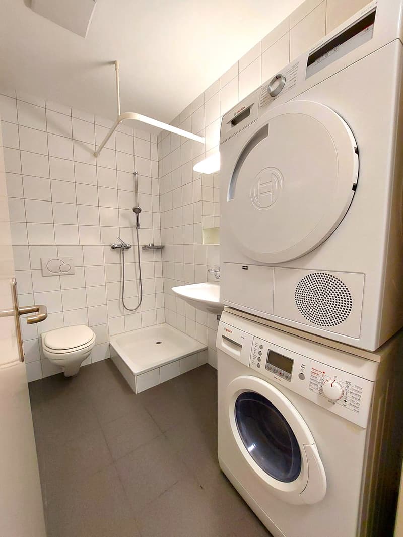 white tiled walls, white tiled floor, toilet, shower, sink, washing machine on top of dishwasher