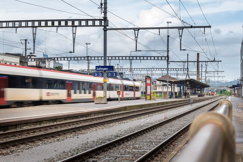 Train station with multiple tracks, train arriving, platform, overhead electrical wires, people waiting
