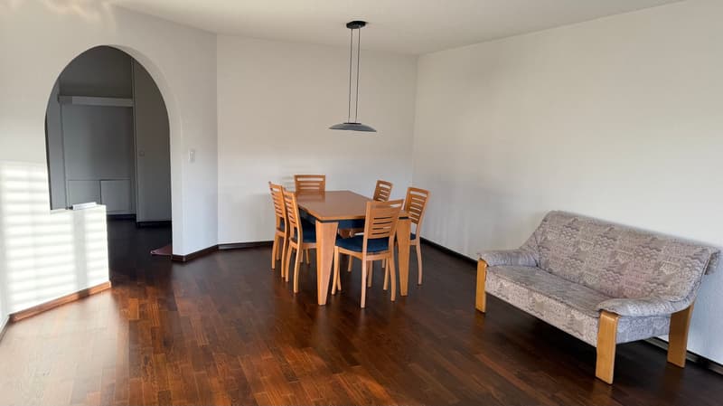 A room with a dining table and 6 chairs, a couch, hardwood floor, white walls, and a pendant light.