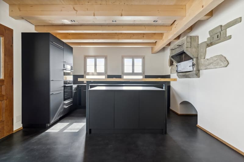 Black cabinets, concrete floor, island with gas stove, microwave and oven, wooden ceiling beams, white walls, stone fireplace