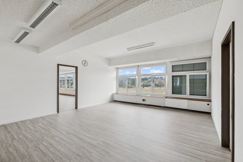 An empty room with white walls, wooden flooring, large windows with shutters, recessed lighting fixtures on the ceiling, a clock mounted on the wall above the window sill, and a mirrored door leading to another room.