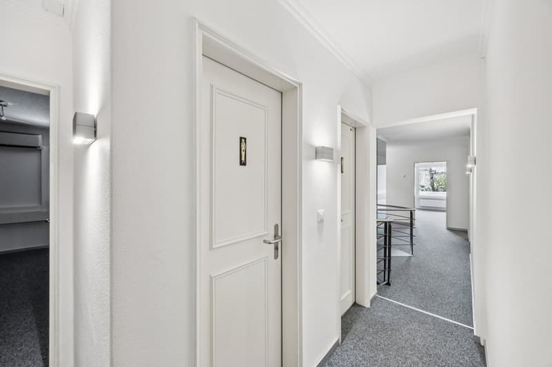 Empty hallway with white walls, gray carpet, white doors, wall-mounted lights, and a staircase.