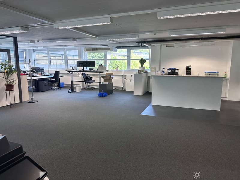 The image shows a modern, open-plan office space with a reception area, desks, computers, and various office equipment. The room has large windows providing natural light, and the walls and floors are in neutral colors. The overall layout and furnishings