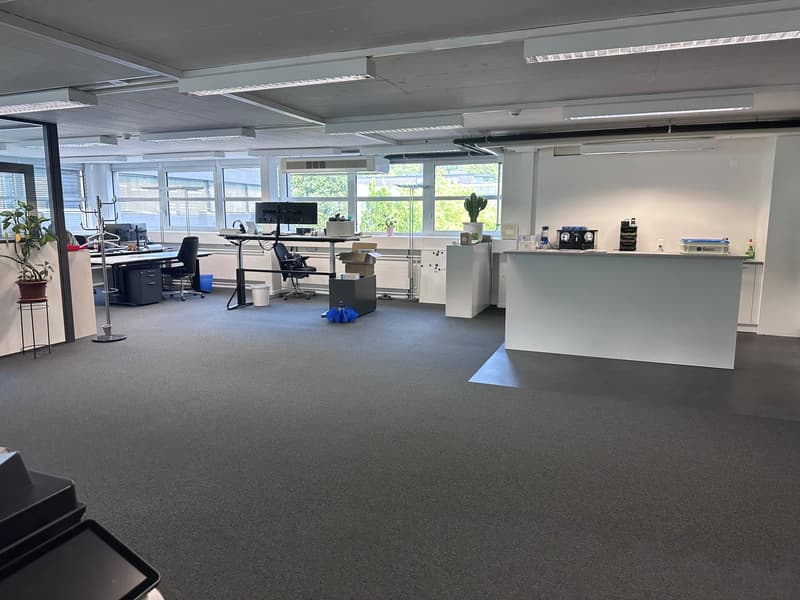 The image shows a modern, open-plan office space with a reception area, desks, computers, and various office equipment. The room has large windows providing natural light, and the walls and floors are in neutral colors. The overall layout and furnishings