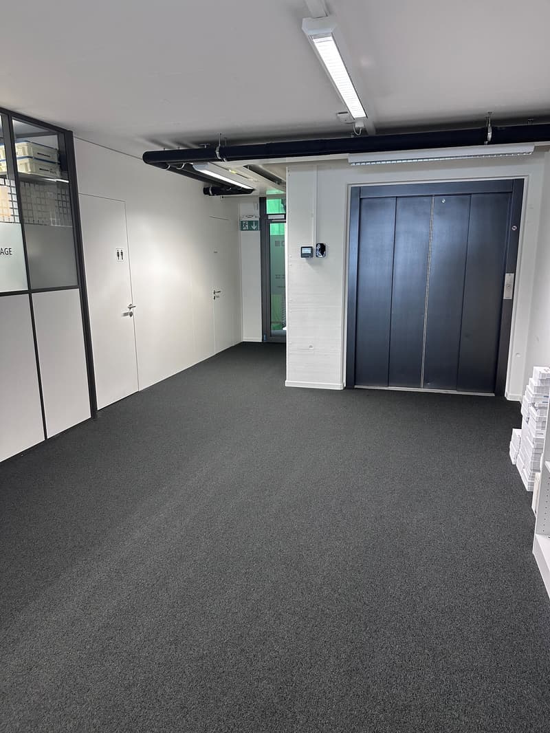 The image shows a spacious office space with a gray carpeted floor, white walls, and a large elevator door. The room is equipped with overhead lighting fixtures and appears to be a commercial or industrial setting.