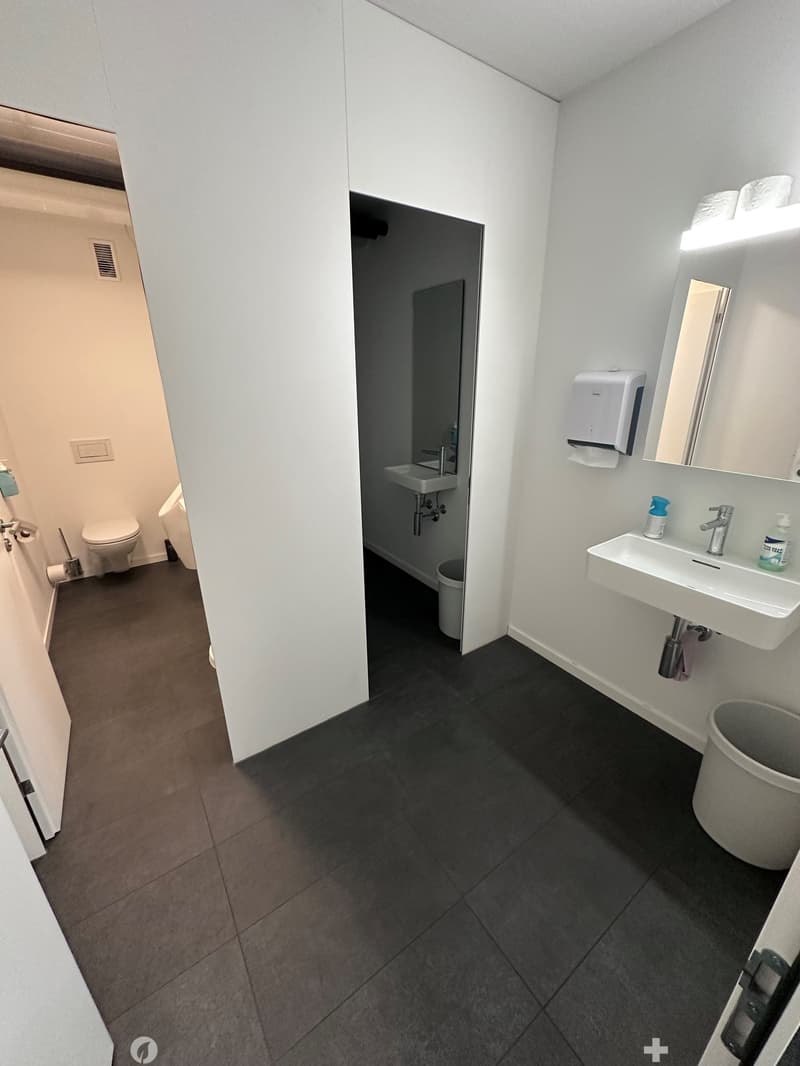 The image shows a bathroom with a white sink, toilet, and a door leading to another room. The floor is covered with dark gray tiles, and the walls are painted white. The bathroom appears to be clean and well-maintained.