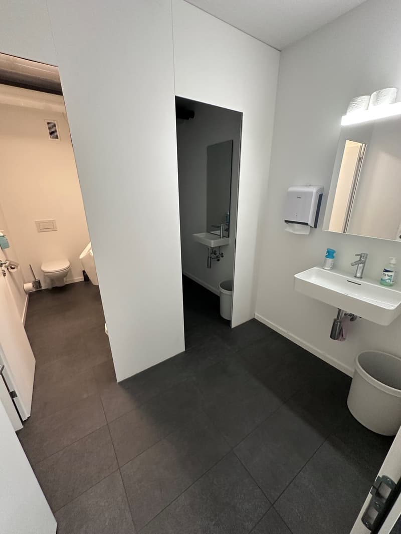 The image shows a bathroom with a white sink, toilet, and a door leading to another room. The floor is covered with dark gray tiles, and the walls are painted white. The bathroom appears to be clean and well-maintained.