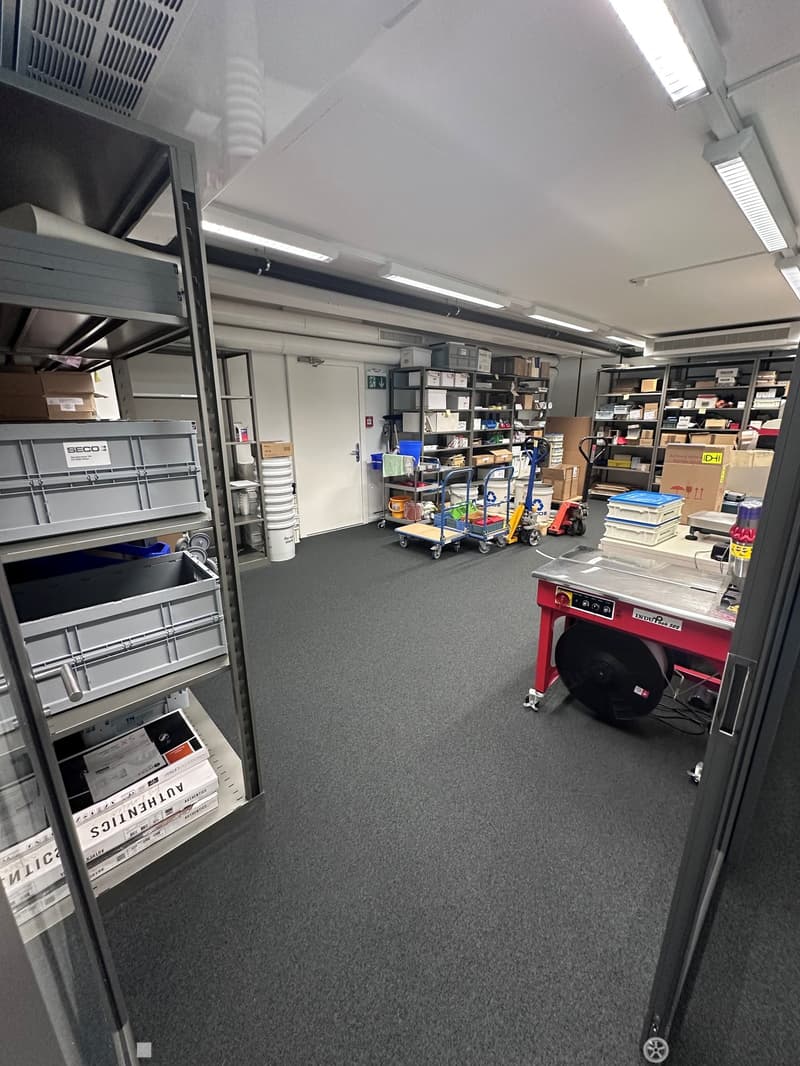 The image shows a storage room or warehouse-like space. It has shelves and storage containers, as well as various equipment and tools. The room has a concrete floor and a high ceiling with lighting fixtures. There appears to be a garage door or entrance v