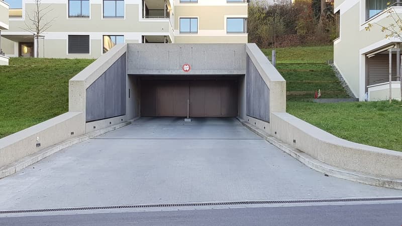The image shows the entrance to an underground garage. The entrance is a large, concrete structure with a sloped driveway leading down into the garage. The garage doors are closed, and there is a red sign on the wall above the entrance.