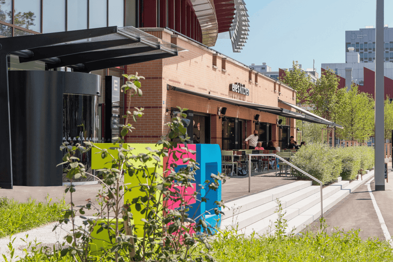 A two-story brick building with a covered outdoor seating area, surrounded by greenery and trees. The building has large windows and a colorful mural on the exterior. There are people visible inside the building, suggesting it is a commercial or public sp