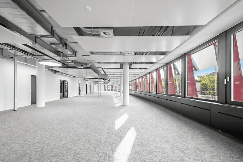 The image shows a large, open commercial space with a long hallway. The space has a modern, industrial design with exposed ceiling beams and ductwork. The walls are white, and the floor is covered in gray carpeting. Large windows line one side of the hall