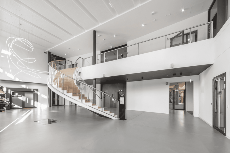 Large open-concept space with high ceilings, modern glass and steel staircase, white walls, and concrete flooring. The space appears to be a commercial or office building with multiple levels and glass doors leading to other areas.