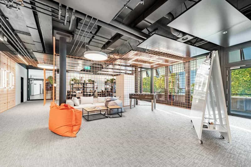 Open concept office space with exposed ceiling, concrete floors, and large windows. The space features a lounge area with an orange bean bag chair, a coffee table, and shelving units. There is also a desk with a computer setup in the background.