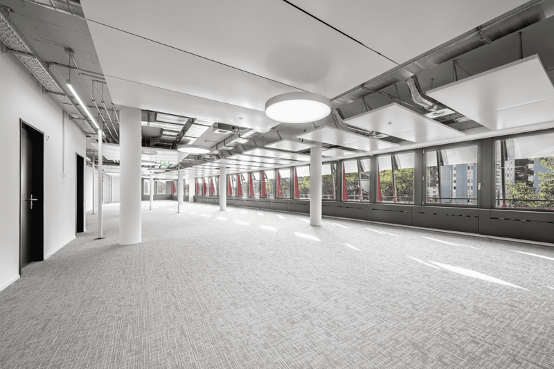 Large open office space with high ceilings, exposed ductwork, and large windows providing natural light. The space has a modern, industrial aesthetic with a gray carpet floor and white walls. There are multiple columns supporting the ceiling and a door vi