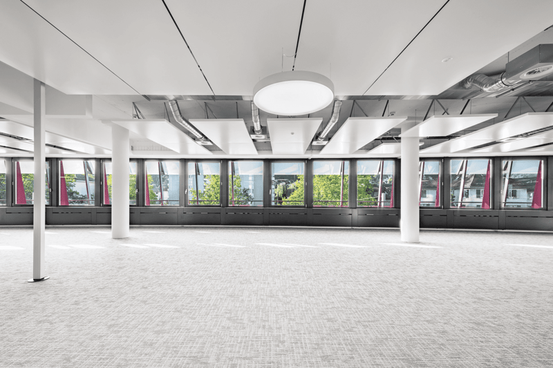 Large open office space with high ceilings, exposed ductwork, and floor-to-ceiling windows providing a view of the surrounding area. The space has a modern, industrial aesthetic with a neutral color palette and concrete flooring.