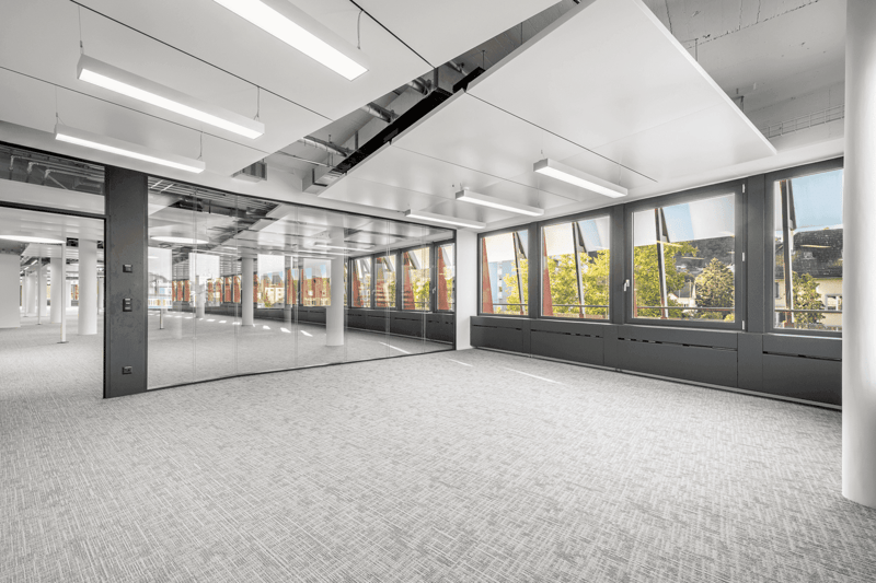 Large open office space with floor-to-ceiling windows, providing a view of the surrounding area. The space has a modern, minimalist design with gray carpeting, white walls, and recessed lighting. There are glass partitions dividing the space, creating a s