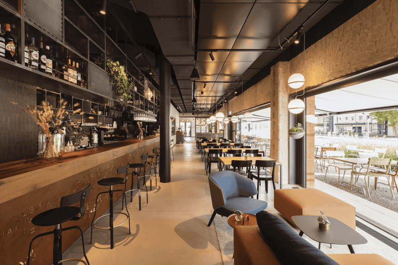 This image shows a modern and stylish cafe or restaurant interior. The space features a long bar area with shelves displaying various bottles and decor. The seating area has a mix of tables, chairs, and couches, creating a comfortable and inviting atmosph