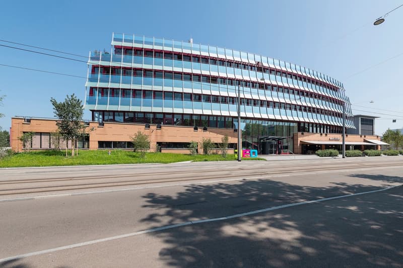 A multi-story office or commercial building with a curved, glass facade and red accents. The building has a modern, architectural design and is surrounded by trees and a paved area in front.