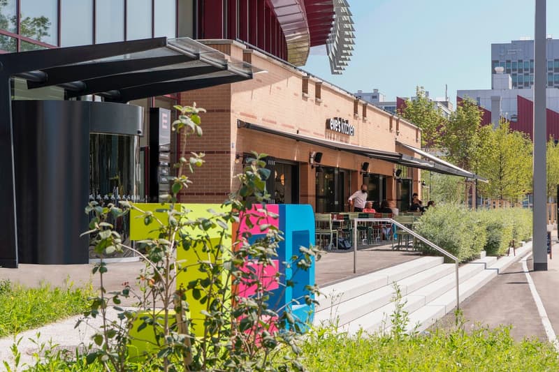 Brick building with a restaurant called 'Gus's Kitchen', outdoor seating area, colorful planters, and a modern architectural design with a canopy structure.