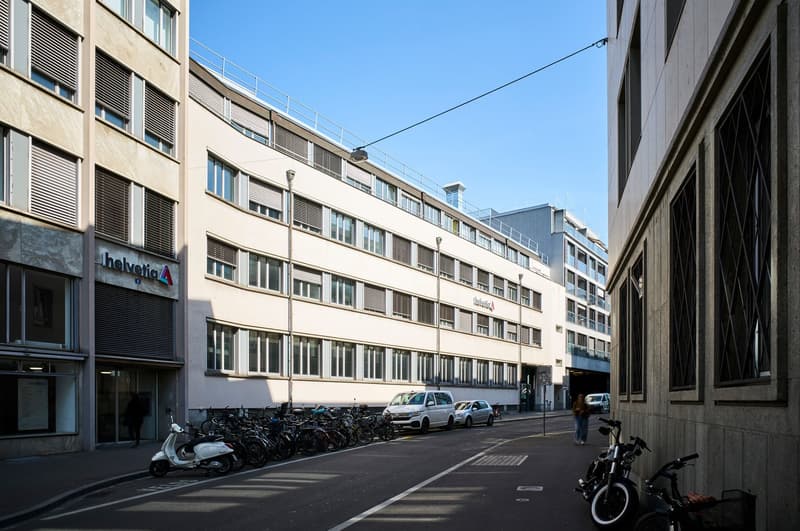 Modern office building with glass windows, multiple floors, and a sign reading 'Helvetic'. Street with parked cars and bicycles, and a pedestrian walking.