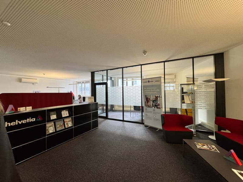 modern office space with glass walls, black and red furniture, carpeted floor, books on a shelf, a table with magazines, and a banner